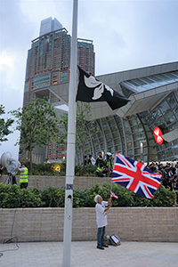Black Bauhinia flag on a flagpole, with 'Grandma Wong' waving a British flag behind, West Kowloon Station, 7 July 2019